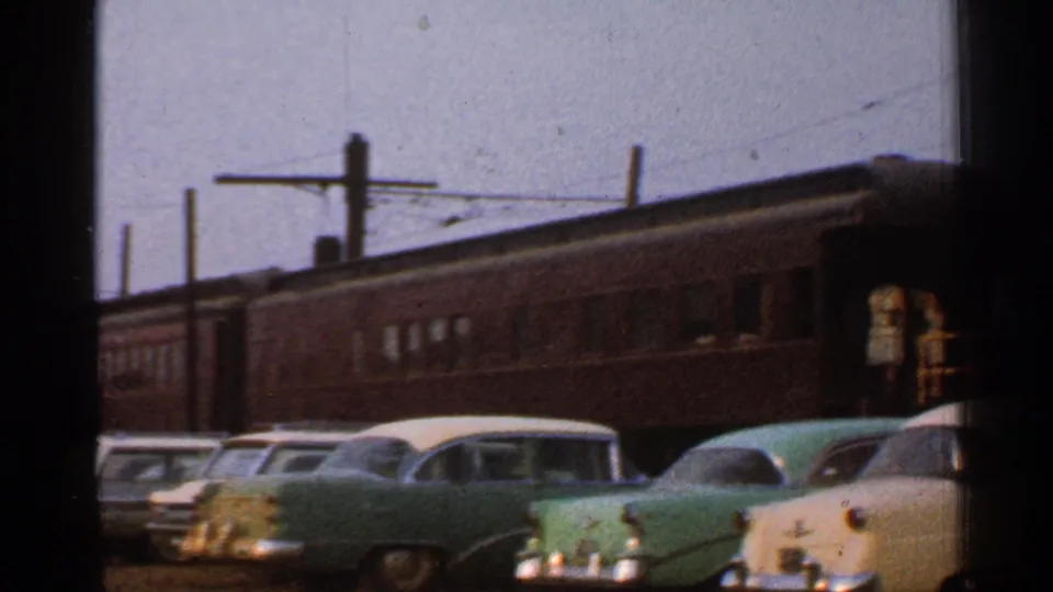 Brown passenger cars and parked 1950s automobiles are visible, with a small person standing near the right edge.