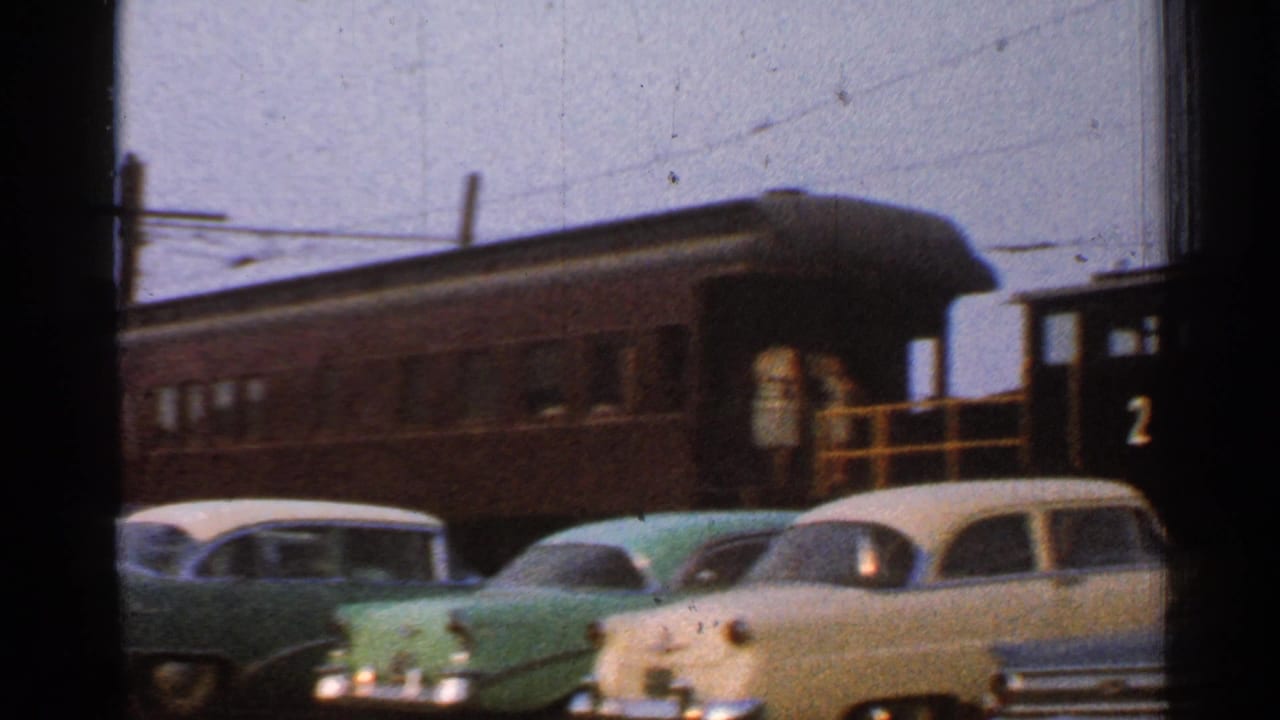 Classic cars parked beside a dark rail car and locomotive in a train yard.