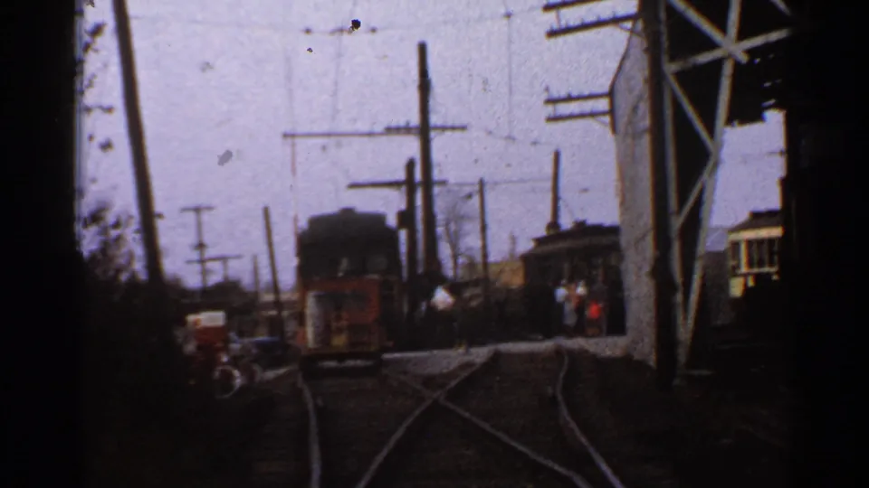 1953 Connecticut frame of an industrial trolley yard with curving rails, overhead electric lines, utility poles, trolley cars, and distant people.