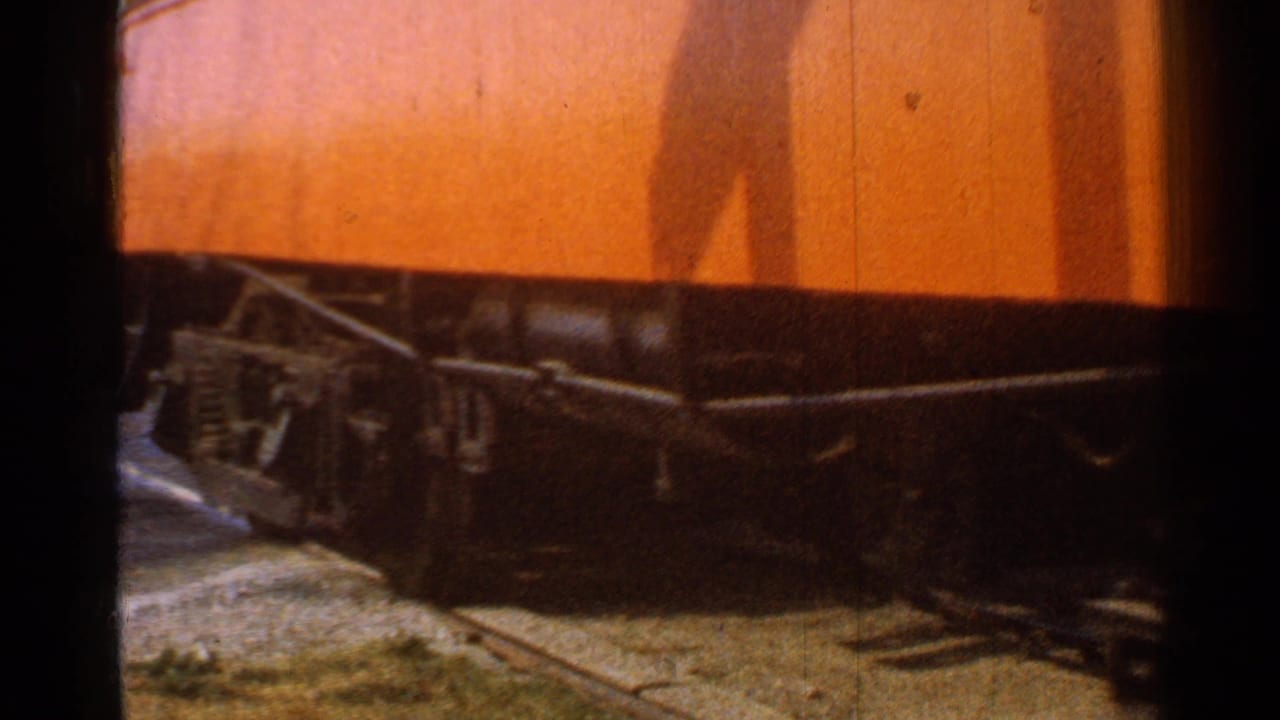 Close 1953 Connecticut rail-yard view of an orange rail car undercarriage and wheels rolling along steel track.
