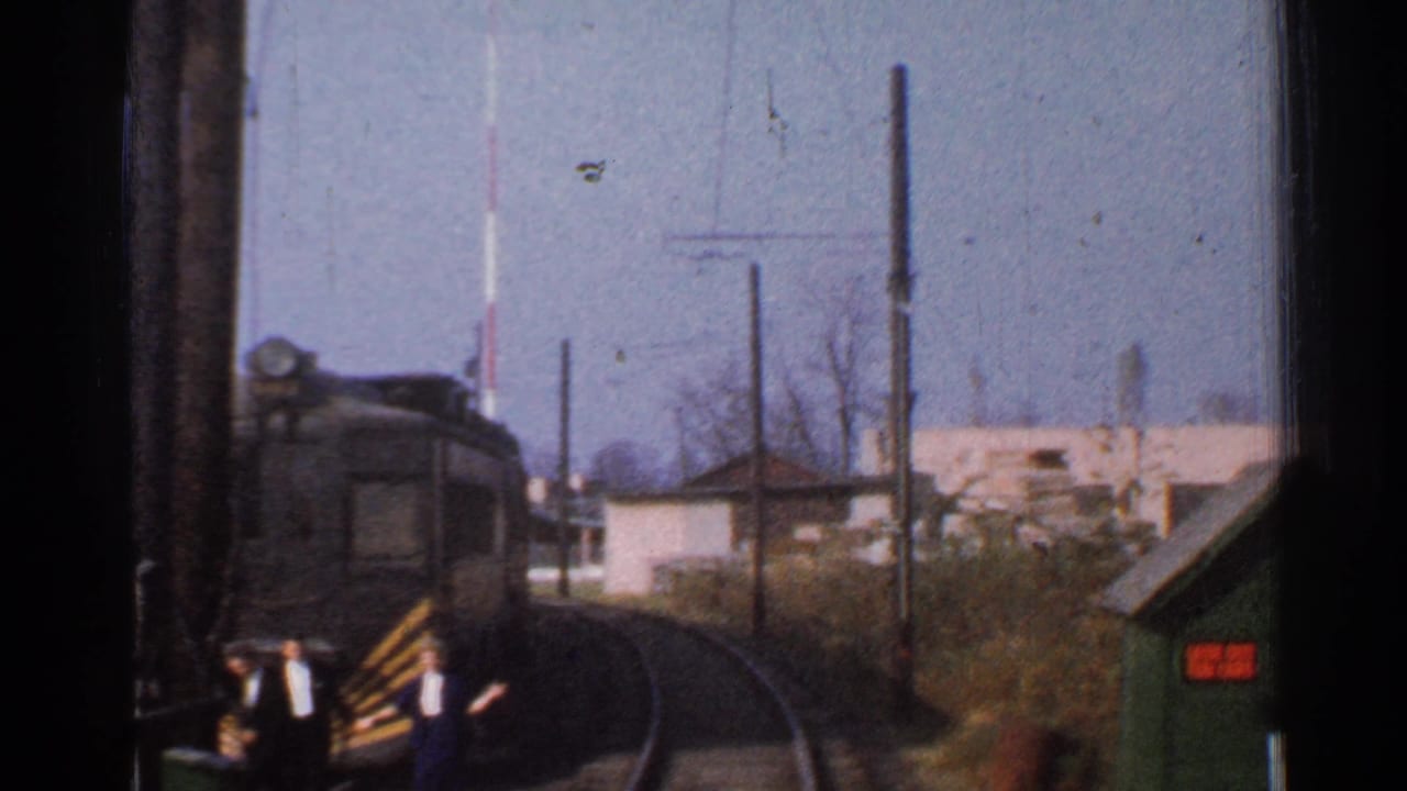 Train moves past a crossing area with people, poles, and low buildings nearby.