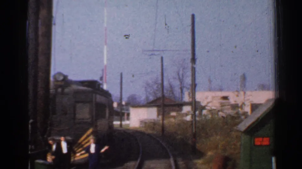 Forward-facing 1953 Connecticut rail view with bystanders near the crossing and trackside buildings beyond.