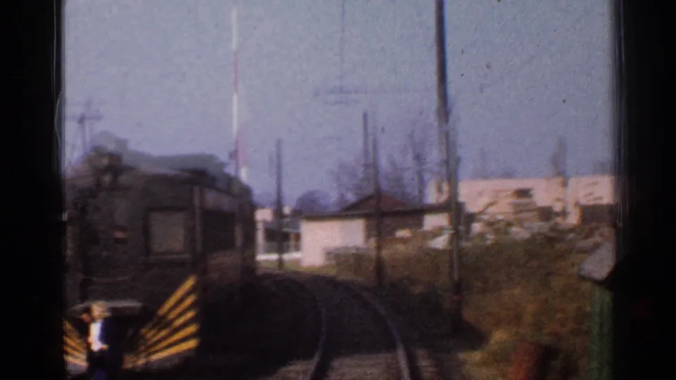 Soft 8mm frame from a moving train in Connecticut, passing a striped gate, utility poles, brush, and low buildings.