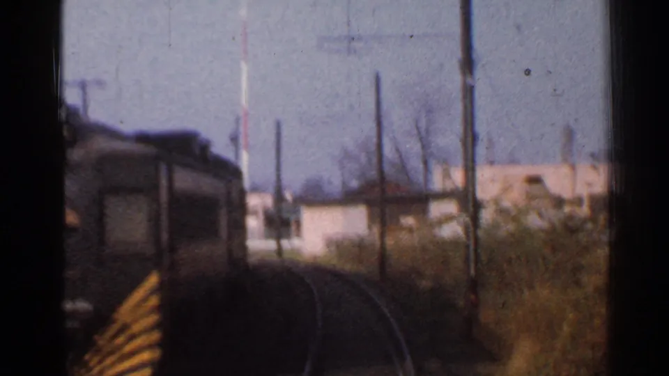 1953 Connecticut rail POV continuing beyond the crossing, with a dark railcar front, poles, brush, and low buildings.
