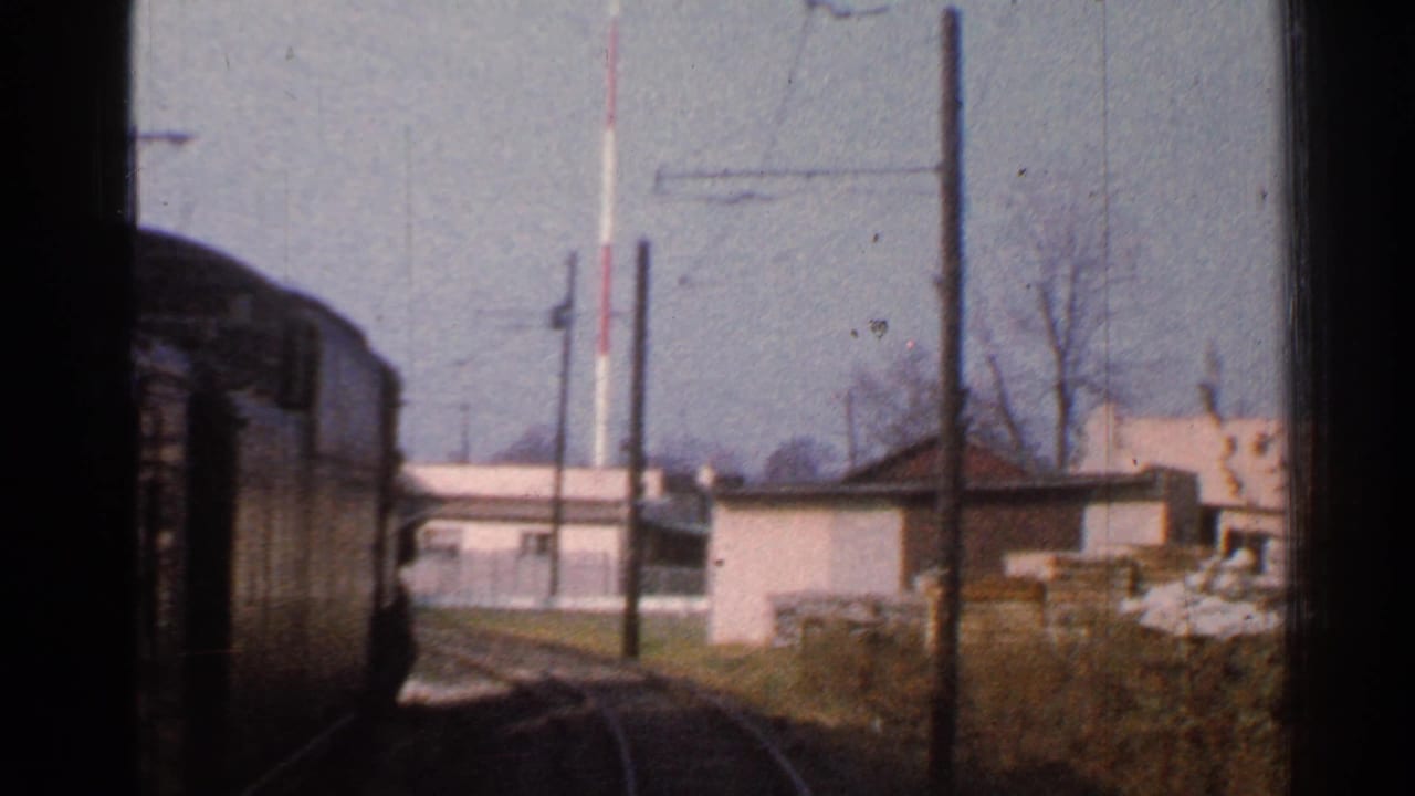 Train-window view of a rail car, low buildings, poles, tracks, grass, and stacked materials near the right side.