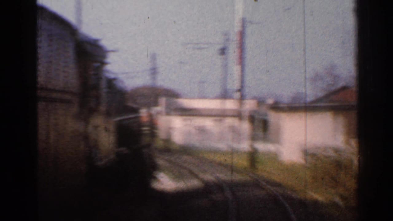 View from a moving train beside curved tracks, small buildings, utility poles, and a dark train edge.