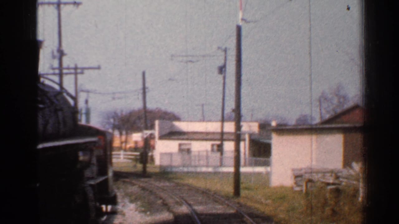 Rail line curves past low buildings, utility poles, fencing, and a red-and-white signal post.