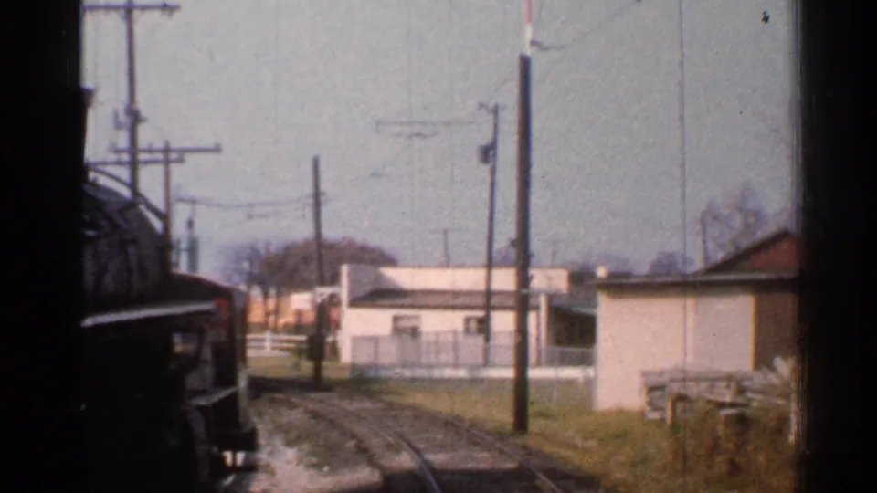 1953 Connecticut train view along curving rails with low trackside buildings, utility poles, fencing, and a red-and-white signal post.