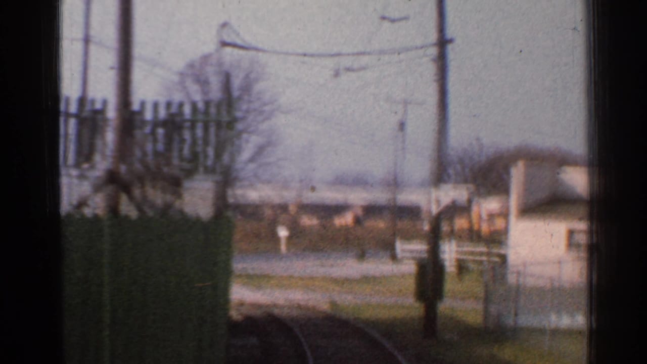 Railway track curves beside a fence, poles, wires, and a small light-colored building.