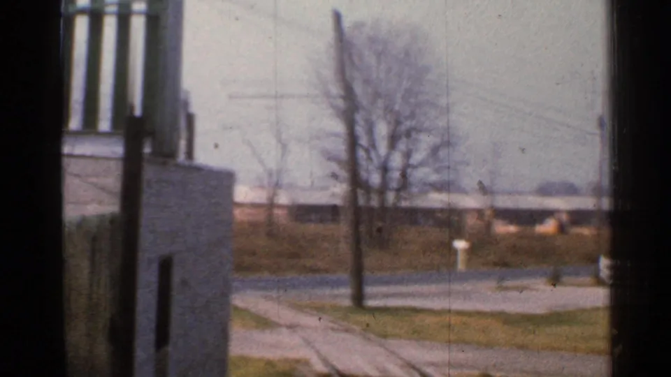 1953 Connecticut rail-side view from a moving train, showing a nearby building edge, road crossing area, poles, and winter-bare trees.