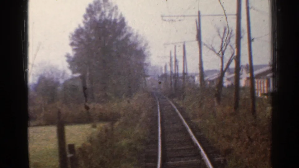 1953 Connecticut home-movie frame showing tracks curving past brush, utility poles, a large tree, and nearby houses from a train window.