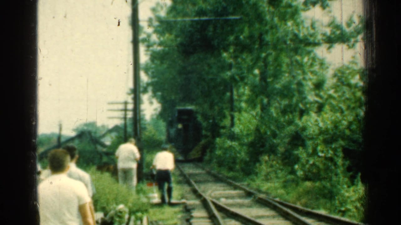 Men in white shirts stand and walk near railroad tracks as a dark rail vehicle approaches.