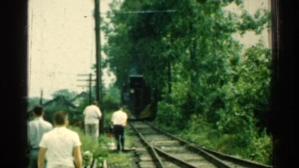 Home-movie frame from 1953 Connecticut showing men from behind near railroad tracks, with trees and an approaching dark rail vehicle.