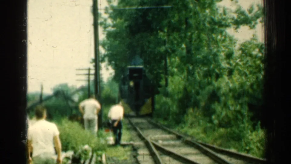 1953 Connecticut 8mm frame of men near curving railroad tracks, dense greenery, utility poles, and a dark rail vehicle with yellow detail.