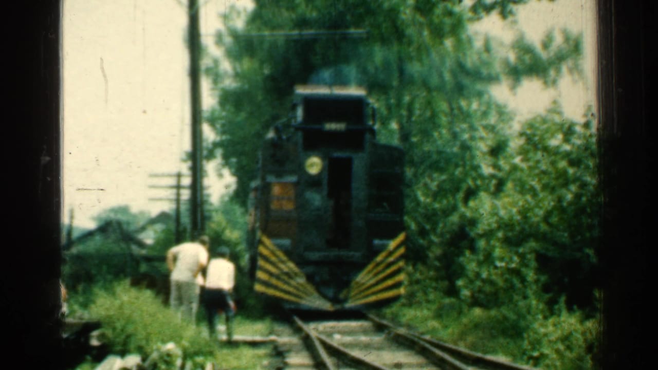 The approaching train is closer in view, with two men near the rails and dense green trees along the track.