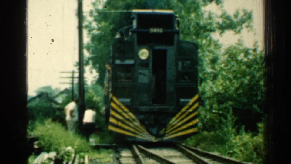 A dark green train with diagonal striping fills more of the frame as two men stand low beside the track in Connecticut footage.