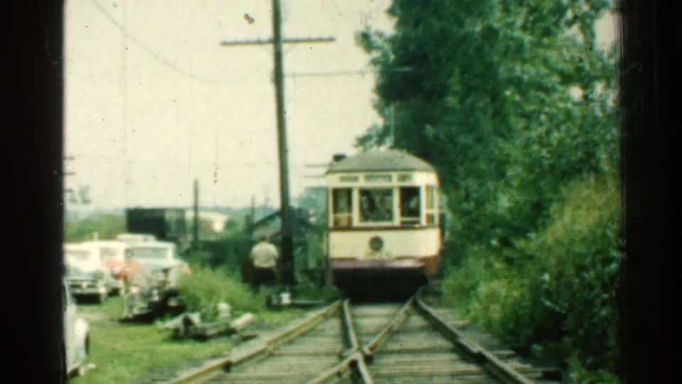 The Connecticut trolley continues toward the camera through a wooded roadside area, with mid-century cars parked along the grass.