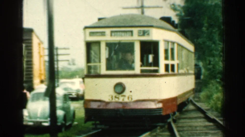 Close 1953 Connecticut frame of trolley number 3876 approaching on rails, with cars and wooded growth along the trackside.