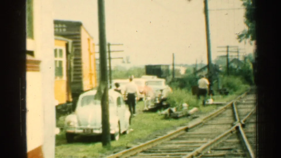 1953 Connecticut trackside frame showing the trolley at left, parked mid-century cars, pedestrians, rails, grass, and utility poles.
