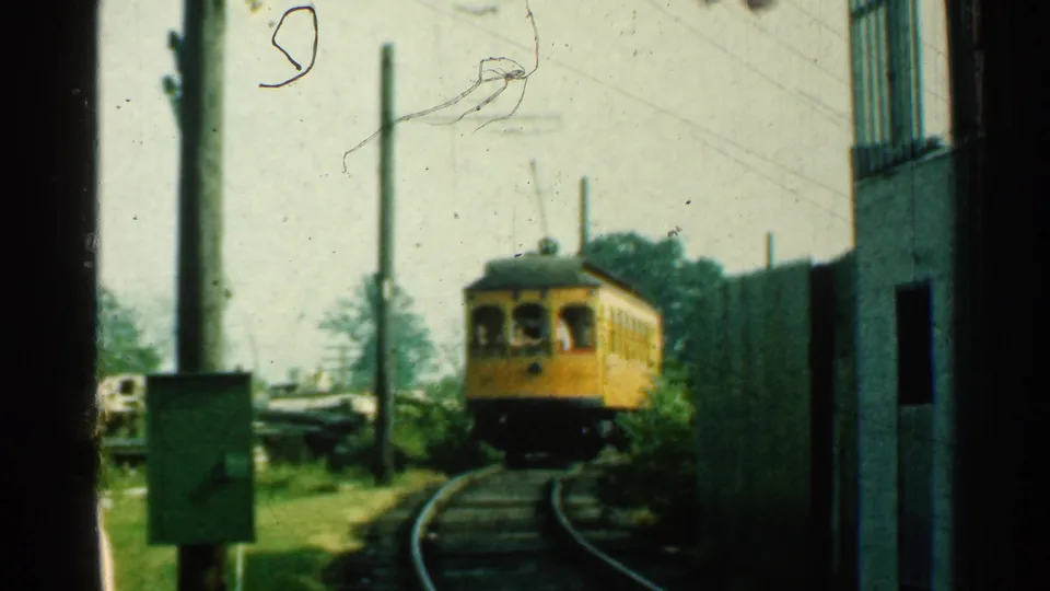 The yellow trolley continues around the bend in Connecticut, framed by trackside poles, grass and a building edge.