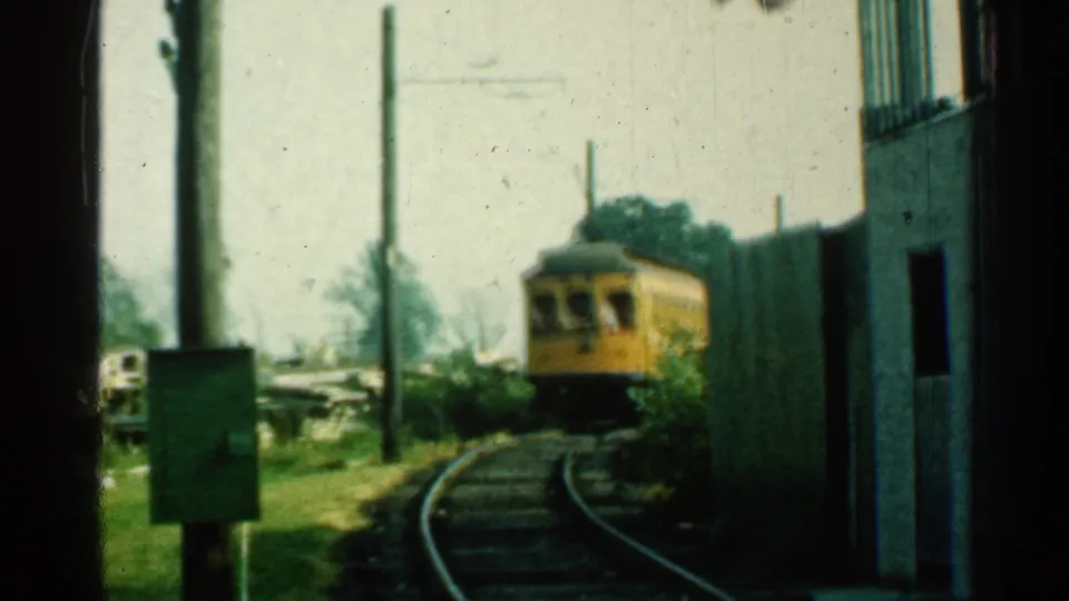 A soft 8mm frame shows the yellow trolley continuing along curved tracks, with poles and a dark doorway framing the view.