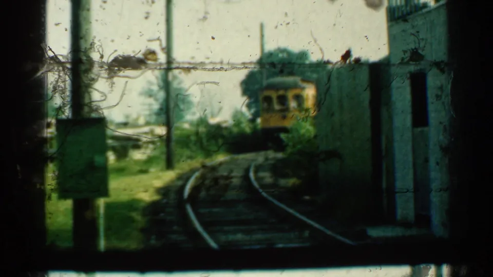 At the end of the clip, the yellow trolley is farther around the bend, with rails, poles, grass and doorway shadows visible.