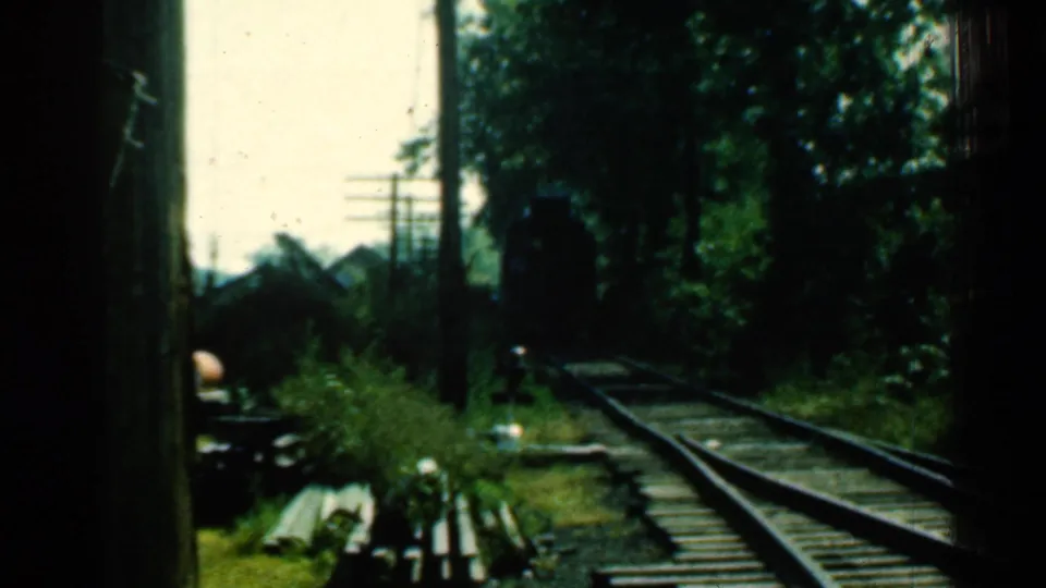 Connecticut railway track scene from the supplied 1953 8mm clip, showing rails, wooden ties, overgrown grass, trees, utility poles, and a shaded foreground edge.