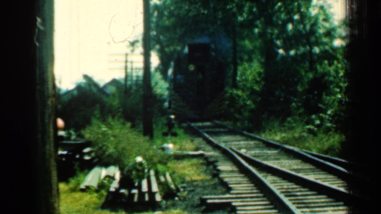1953 Connecticut home-movie frame with an approaching train on tracks framed by trees and dark gate edges.