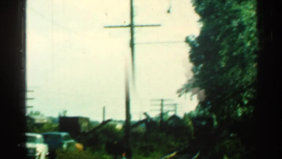1953 Connecticut home-movie frame showing rail tracks, roadside cars, utility poles and trees before the train comes fully into view.