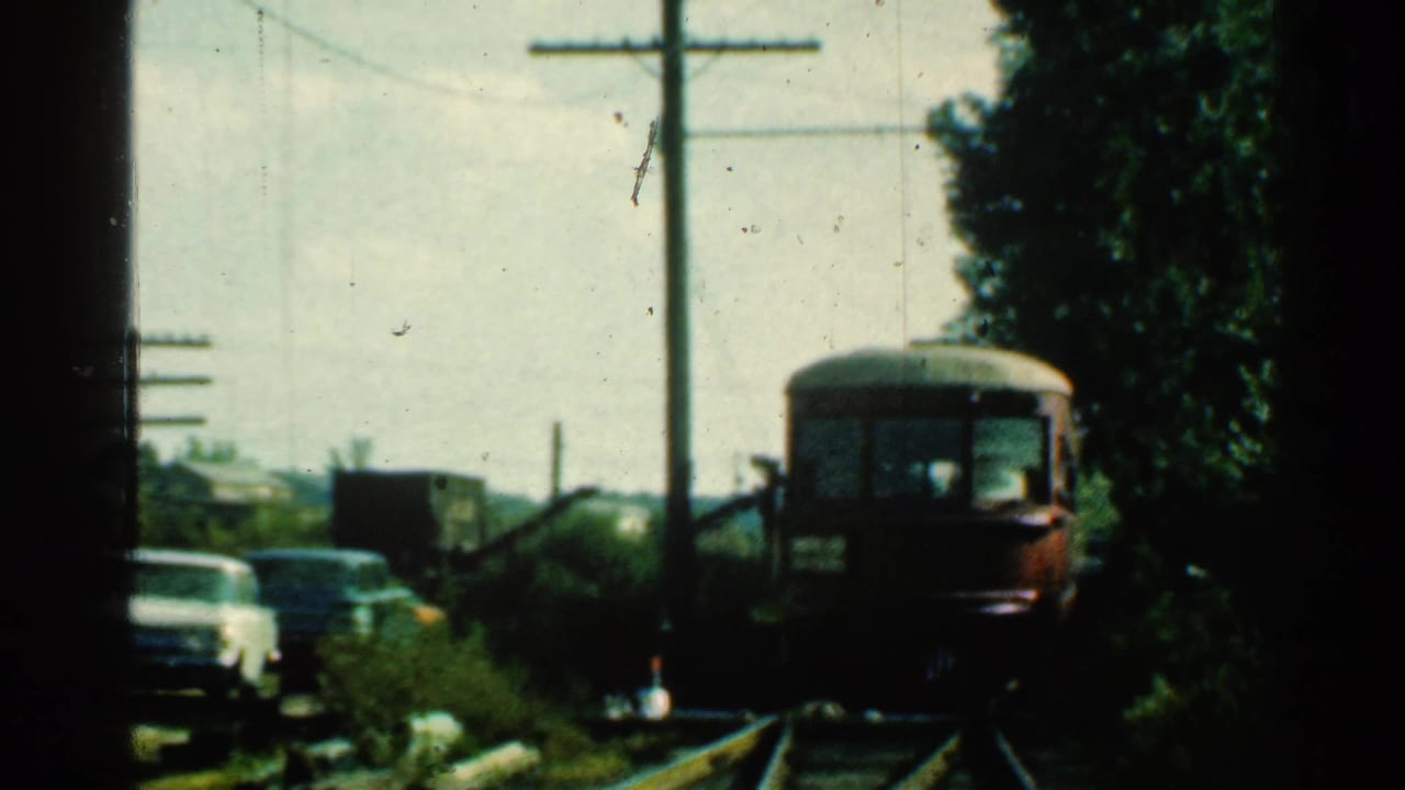 Red train approaching on tracks beside parked mid-century cars and utility poles.