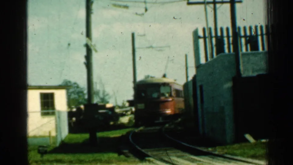 The red trolley moves through the curve on narrow tracks beside a small white building, overhead wires, utility poles, and grass in 1953 Connecticut home-movie footage.