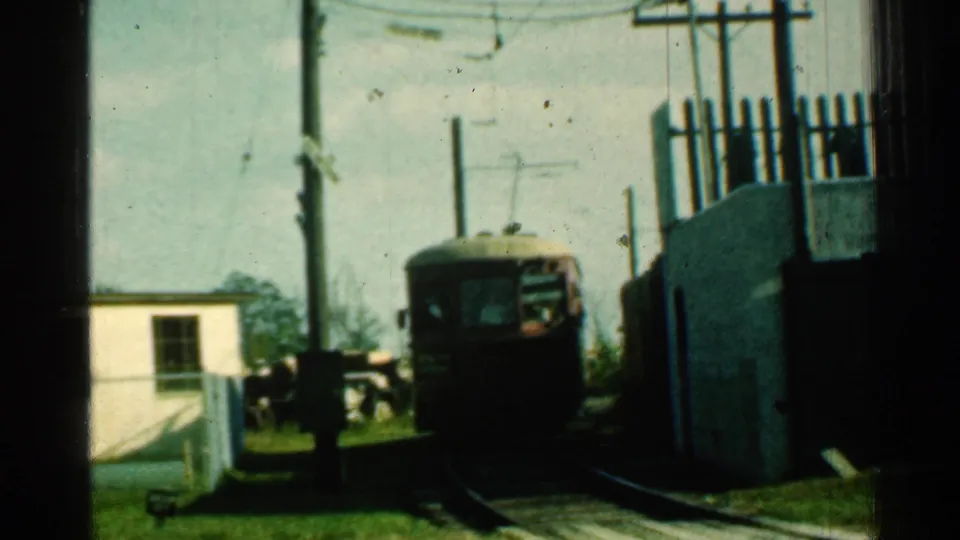 A red trolley faces toward the camera while following the curved electrified track, framed by utility poles, overhead wires, grass, and a small white building.