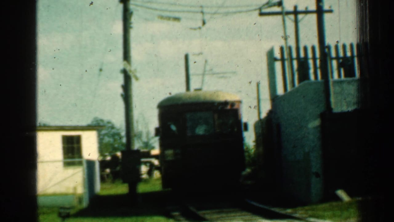 Distant trolley approaching a rural crossing near utility poles and a small white building.
