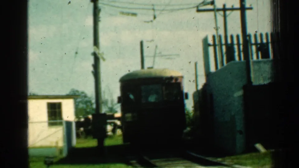 1953 Connecticut 8mm frame showing a trolley approaching a rail crossing with utility poles, wires, grass, and a small white structure nearby.