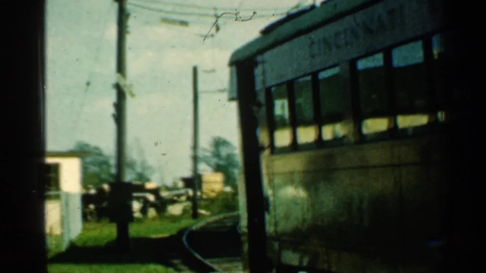 The trolley moves out of frame along a curved track at the rural Connecticut crossing, with poles, wires, grass, and the small white building still visible.