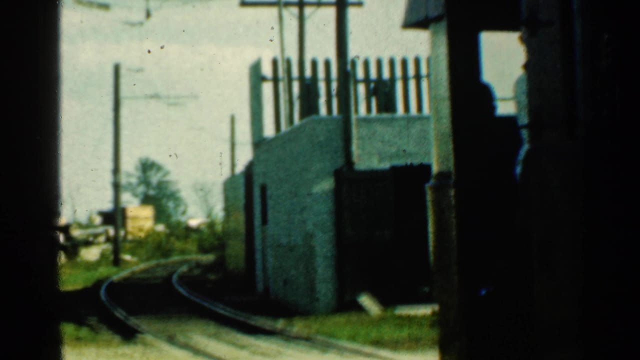 Brown trolley car passes beside curved tracks and a pale roadside structure.