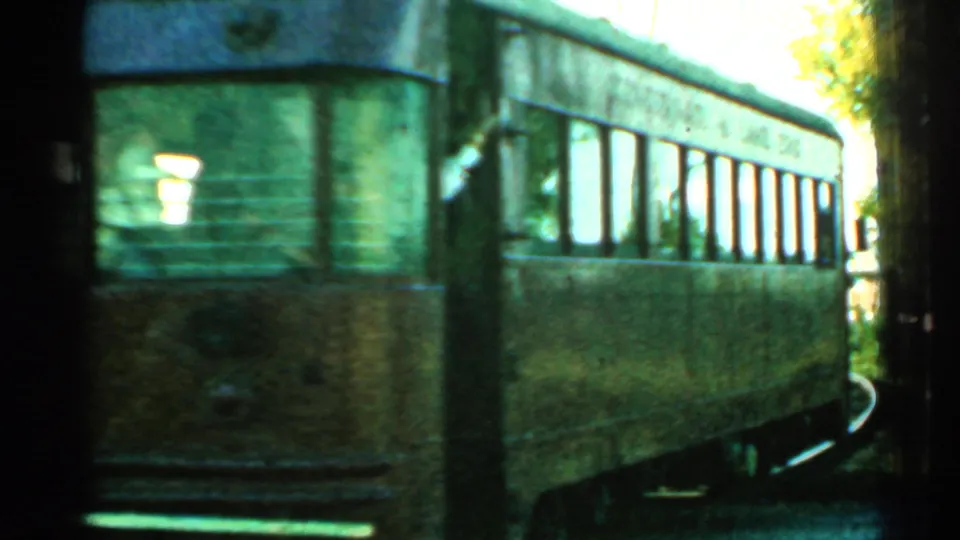 The brown trolley continues around a curve, its windows reflecting light while indistinct people appear inside the car.