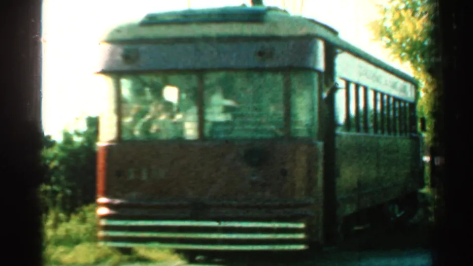 1953 Connecticut 8mm frame showing a brown trolley car near the camera on rails, with trees, brush, and overhead wire nearby.