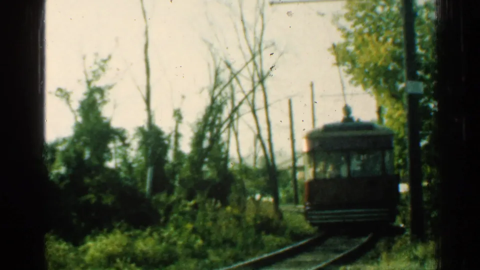 1953 Connecticut 8mm frame showing the trolley farther from the camera on rails, framed by brush, trees, a pole, and pale sky.