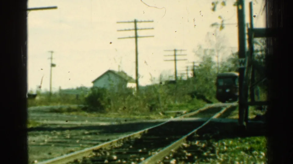 1953 home-movie frame of a train nearing a rural Connecticut crossing, with rails, poles, brush, and a small white house in soft focus.