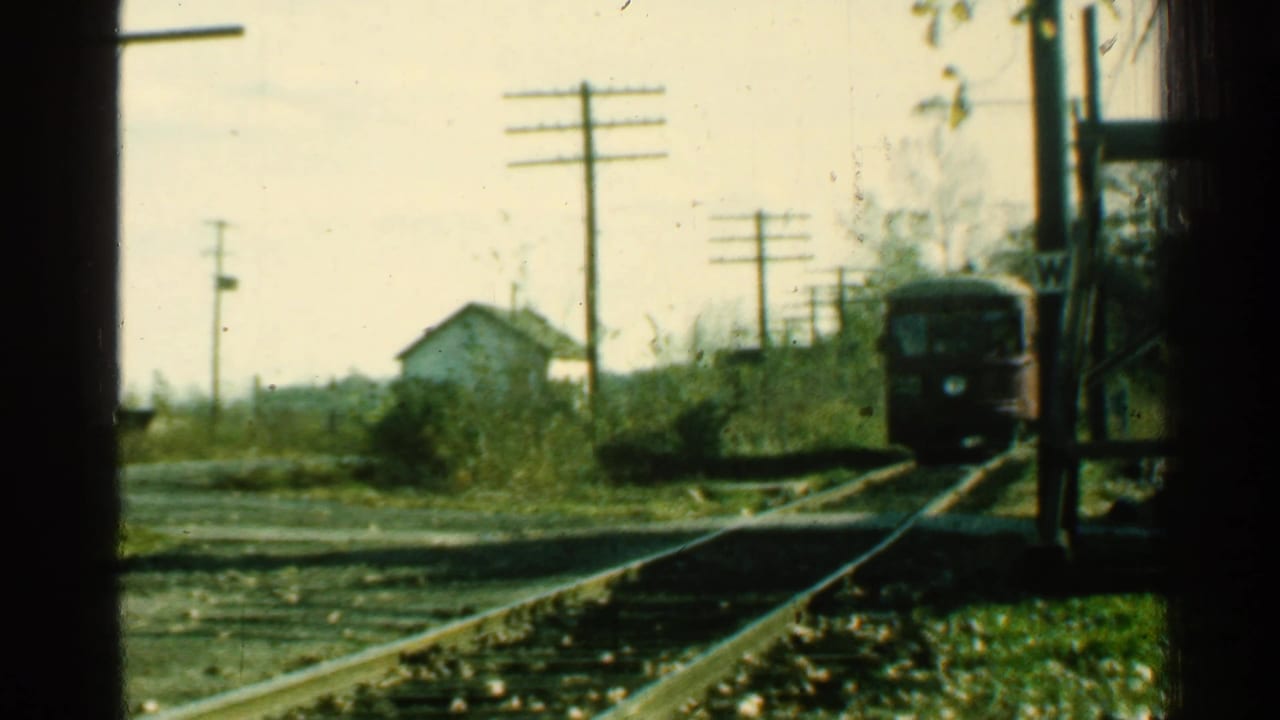 Red trolley in the distance on rural tracks with a small house and utility poles behind it.
