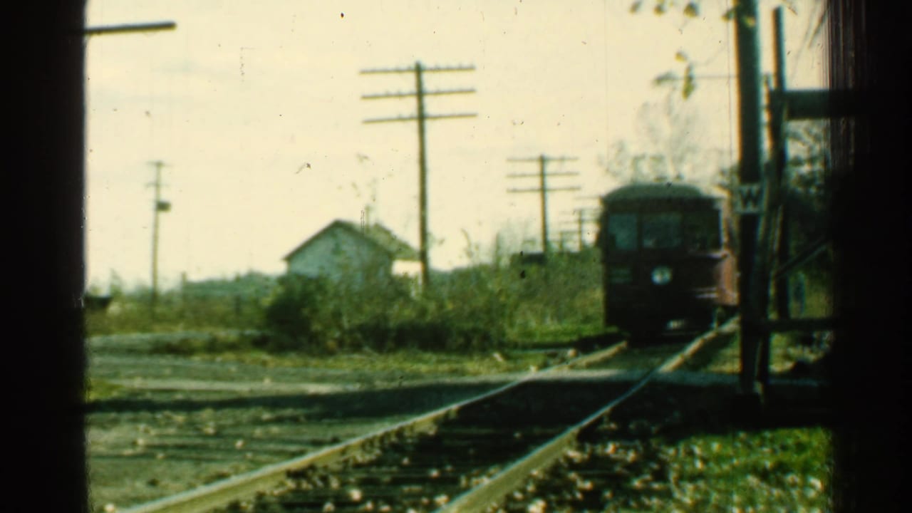 Red trolley closer on the track, seen beside utility poles, brush, and a small house.
