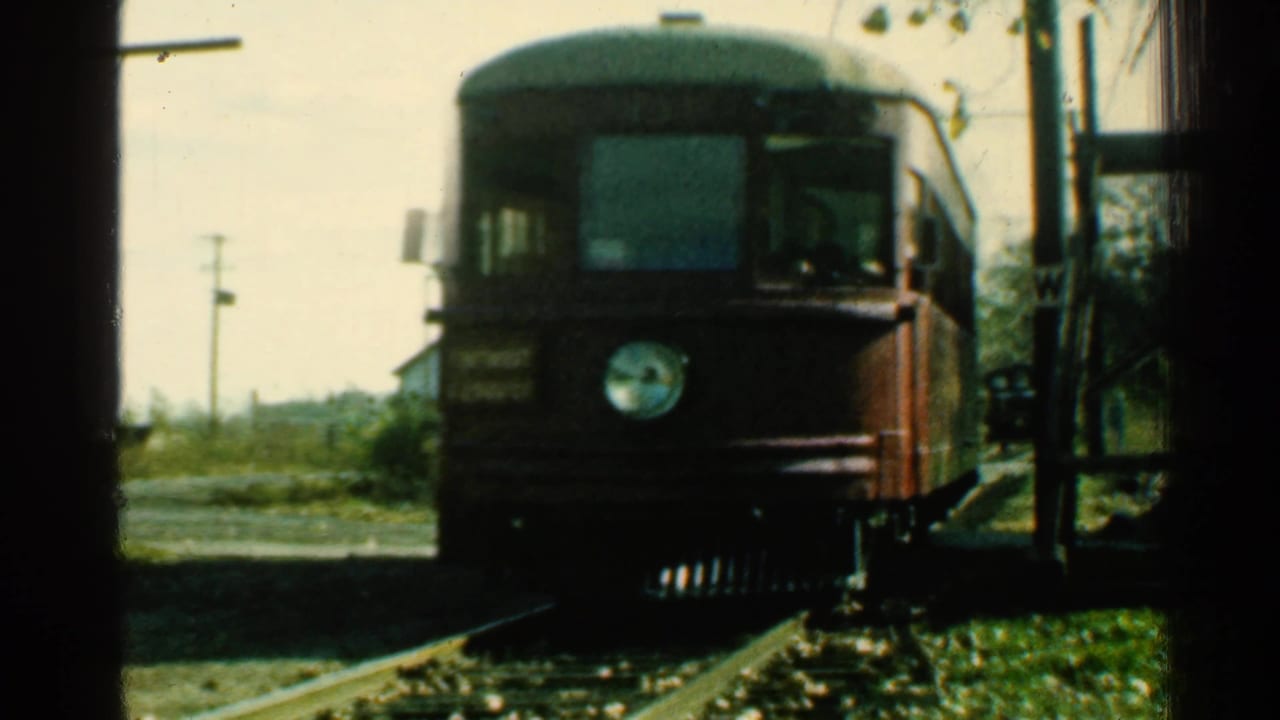 1953 Connecticut 8mm frame showing a red rail car approaching on tracks beside a crossing, utility poles, brush, and a small house area.