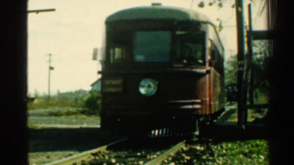 1953 Connecticut 8mm frame showing a red rail car approaching on tracks beside a crossing, utility poles, brush, and a small house area.