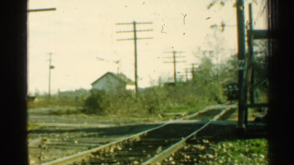 After the rail car has passed, the frame shows empty tracks, a rural crossing, utility poles, brush, and a small light-colored house.