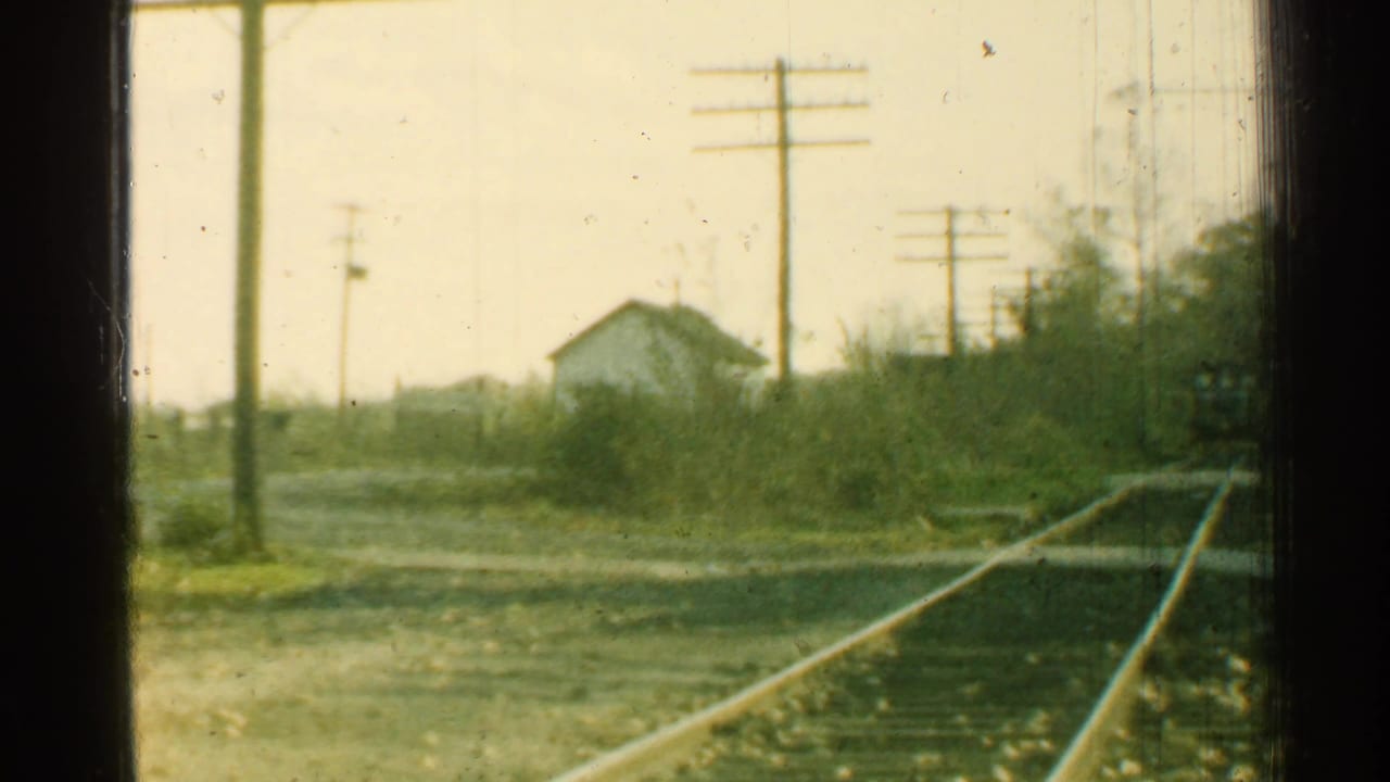 1953 Connecticut home-movie view from a moving train, with tracks, utility poles, brush, and a small white building ahead.
