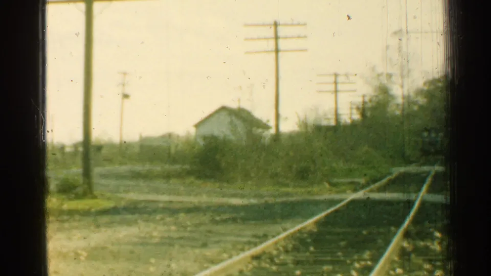 1953 Connecticut home-movie view from a moving train, with tracks, utility poles, brush, and a small white building ahead.