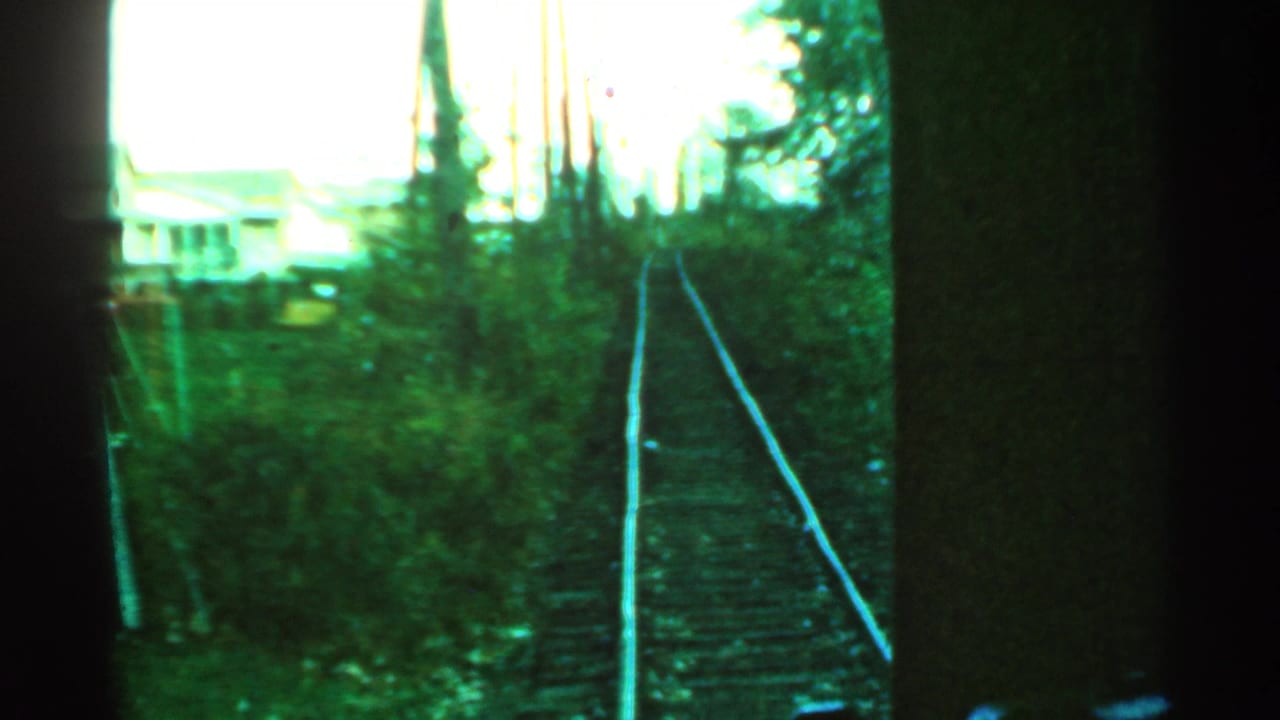 Train tracks viewed ahead through a dark window frame, with trees and a distant house at left.