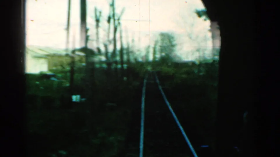 End frame of the 1953 Connecticut train-window view, with tracks, brush, poles, and distant trees softened by motion and film grain.