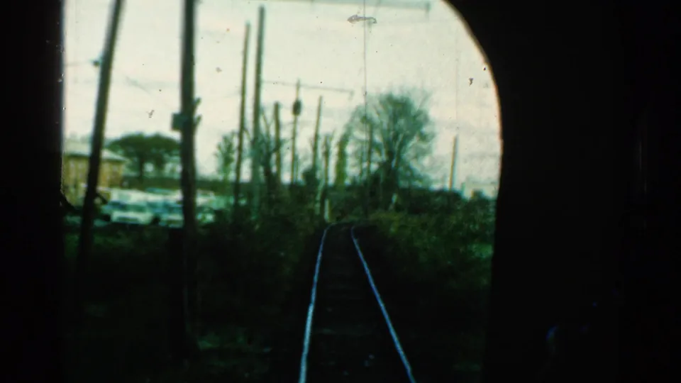 1953 Connecticut home-movie view through a dark train window, showing curving tracks, utility poles, brush, and a cluttered lot beyond.